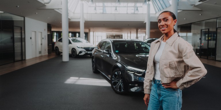 
				Mercedes-Benz Moderatorin Yasmine Blair steht vor einer schwarzen E-Klasse und hlt ihre Hand an der linken Hfte, im Hintergrund parkt ein weier EQE SUV.
				