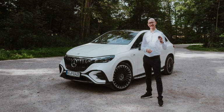 
				Mercedes-Benz Members Produkttrainer Christoph Geissler steht neben einem Mercedes-AMG EQE 43 SUV in MANUFAKTUR opalithwei bright und hlt den Fahrzeugschlssel lchelnd in der Hand.
				