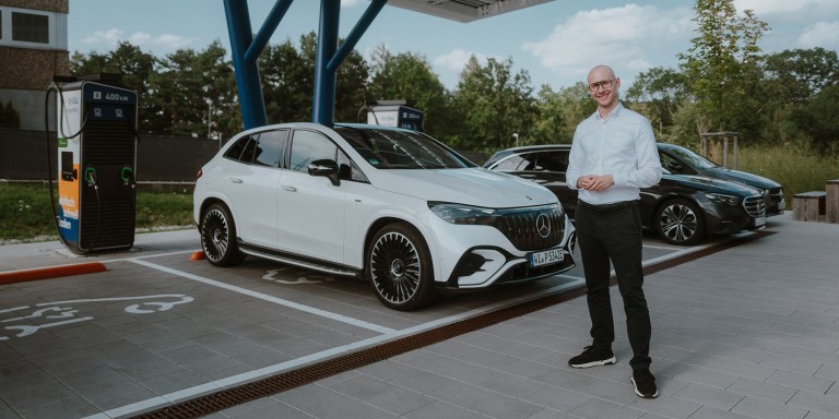 
				Mercedes-Benz Produkttrainer Christoph Geissler steht an einer ffentlichen Ladesule vor einem Mercedes-AMG EQE 43 SUV in MANUFAKTUR opalithwei bright, dahinter parken zwei weitere Mercedes-Benz Fahrzeuge.
				