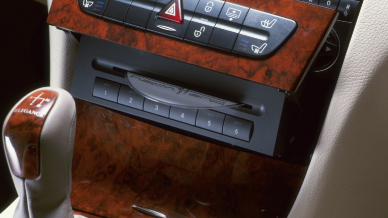
			Blick auf die Mittelkonsole einer E-Klasse Limousine aus dem Jahr 2002 mit Holzoptik und 6-fach-CD-Wechsler hinter der schwenkbaren Schalterleiste, aus dem eine CD ausgeworfen wird.
		